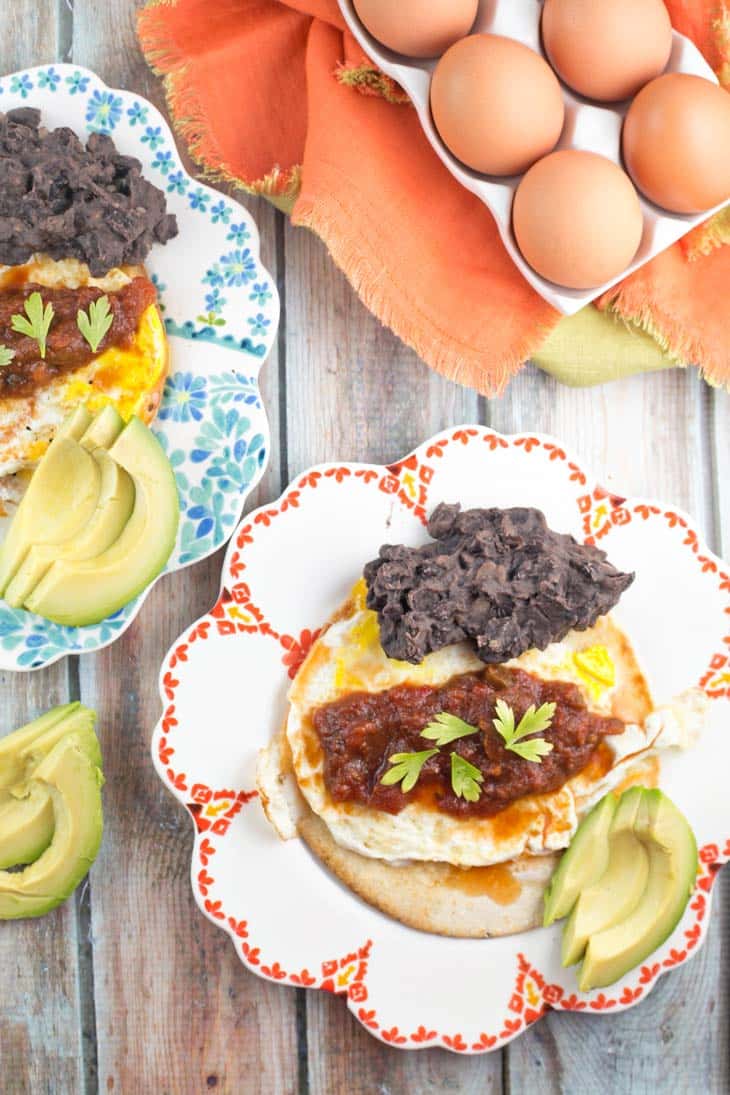 two plates of huevos rancheros surrounded by brown eggs and sliced avocado.