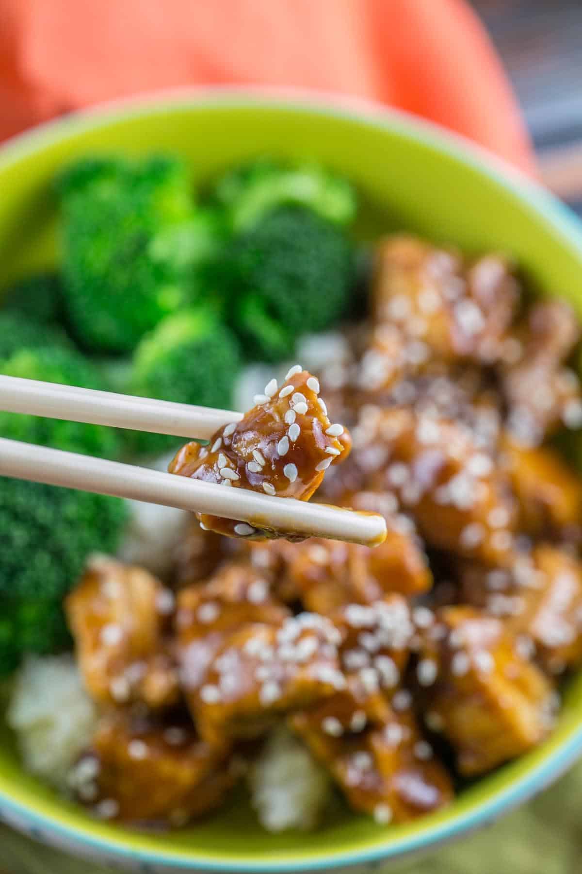 chopsticks holding a bite sized cube of chicken breast covered with sesame seeds