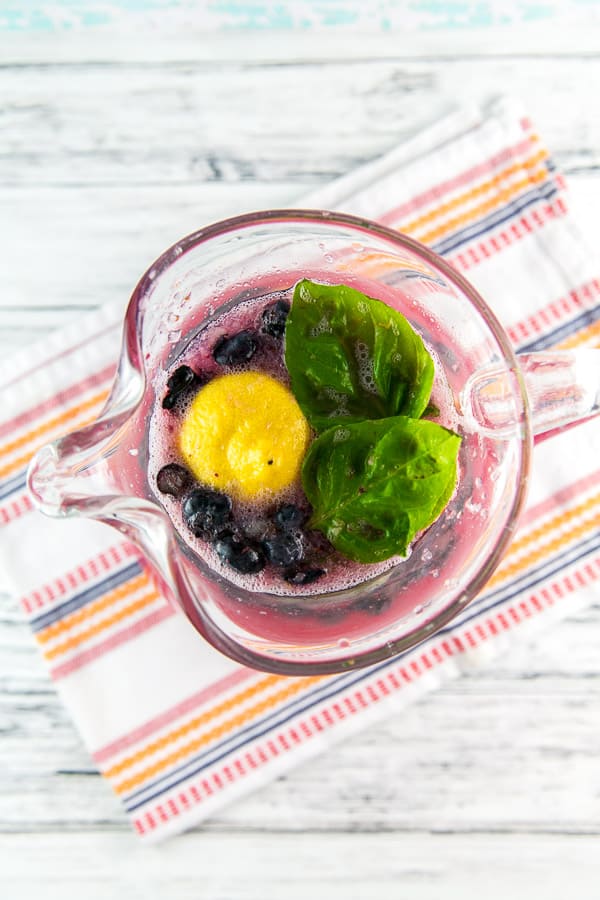 overhead view of a pitcher of blueberry basil lemonade showing a lemon half and two large basil leaves floating on the top of the lemonade