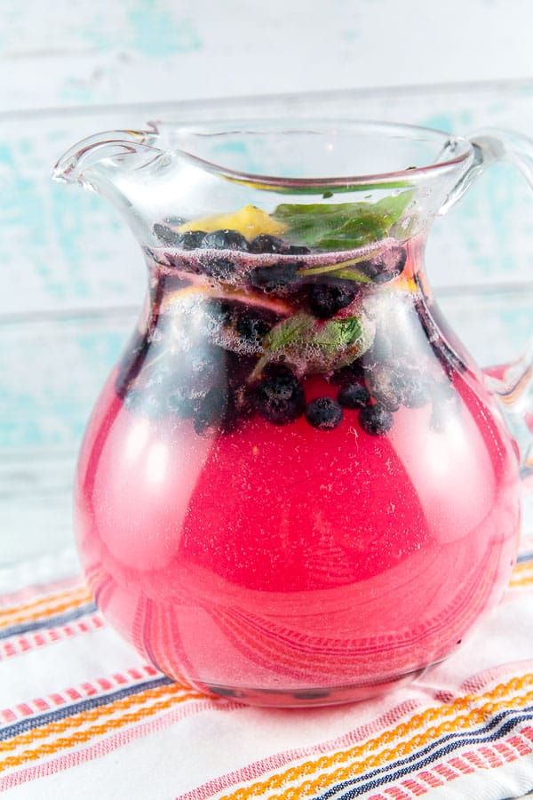 a large glass pitcher filled with blueberry basil lemonade with fresh blueberries, lemon wedges, and leaves of basil floating on the top