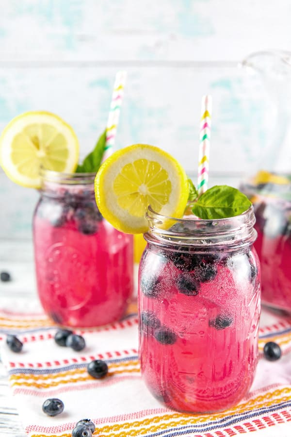 three mason jars filled with blueberry basil bourbon lemonade with wedges of lemon and colorful paper straws