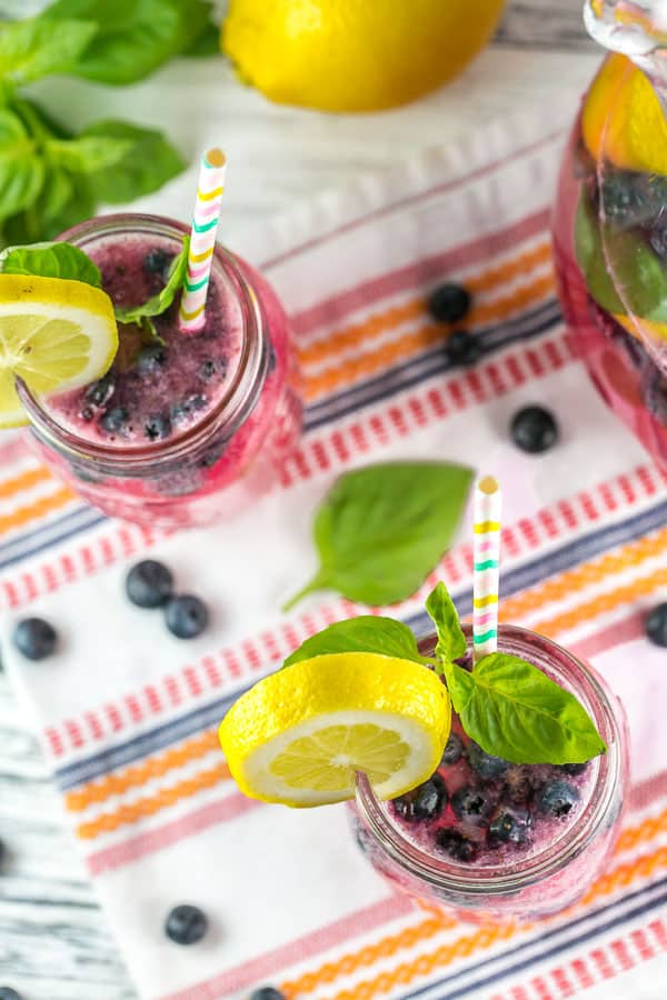 overhead view of mason jars filled with blueberry basil bourbon lemonade on a brightly colored striped dishtowel surrounded by blueberries, basil leaves, and fresh lemons