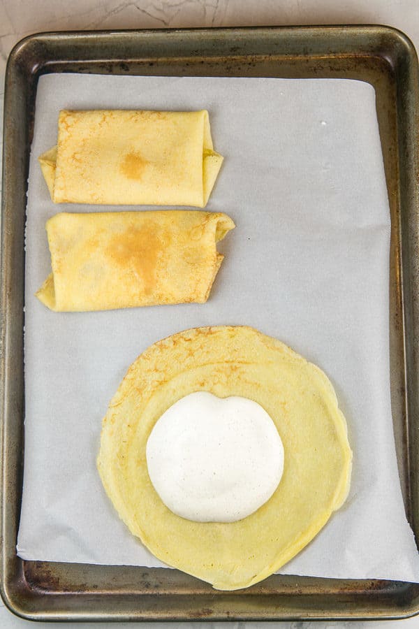 two rolled blintzes on a sheet pan next to a round crepe with a dollop of blintz filling in the center