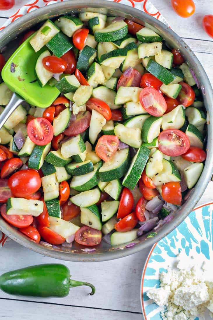 sauteed zucchini, tomatoes, and red onion in a skillet next to a jalapeno pepper and cotija cheese