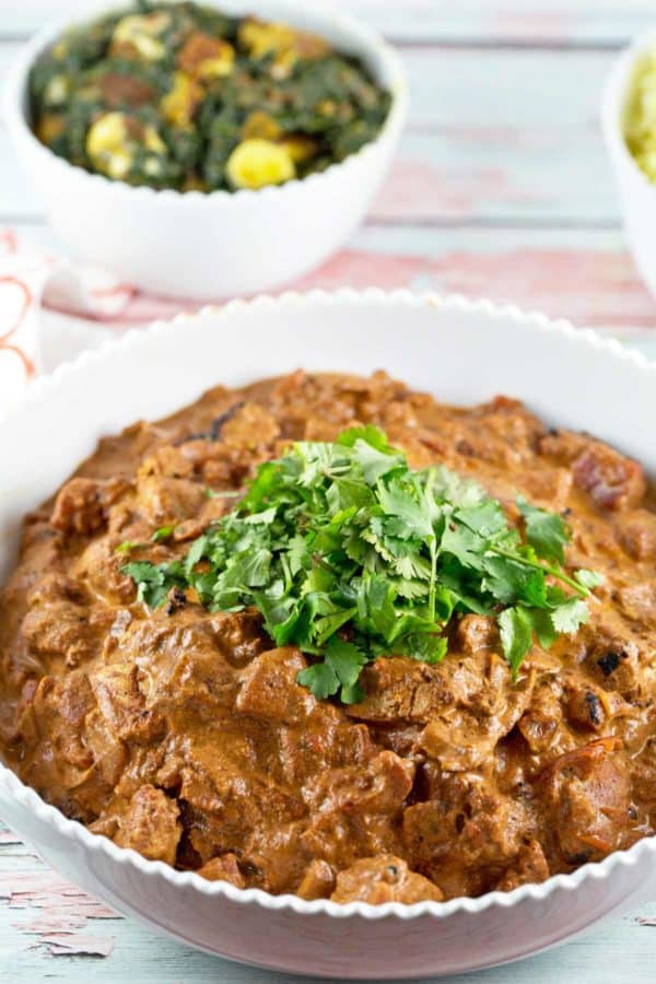 side view of chicken tikka masala in a white serving bowl with saag paneer visible in the background