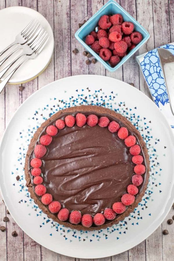 an uncut flourless chocolate cake covered in chocolate ganache on a decorative plate with a basket of raspberries and dessert plates in the background