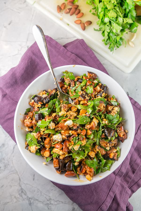 a white serving bowl filled with roasted eggplant and smoked almond dip with a white cutting board with parsley and smoked almonds in the background.