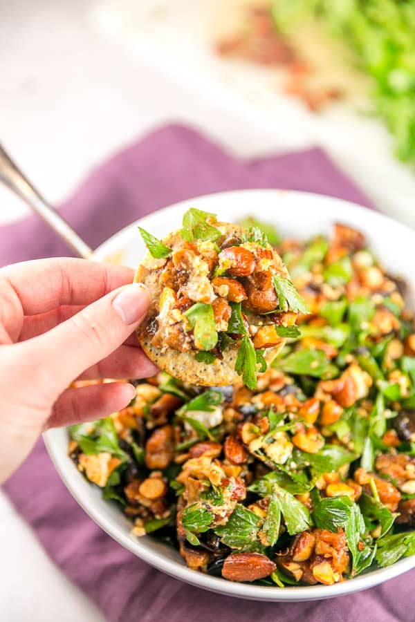 a hand holding a large cracker scooping up some roasted eggplant and smoked almond dip from a large white bowl.