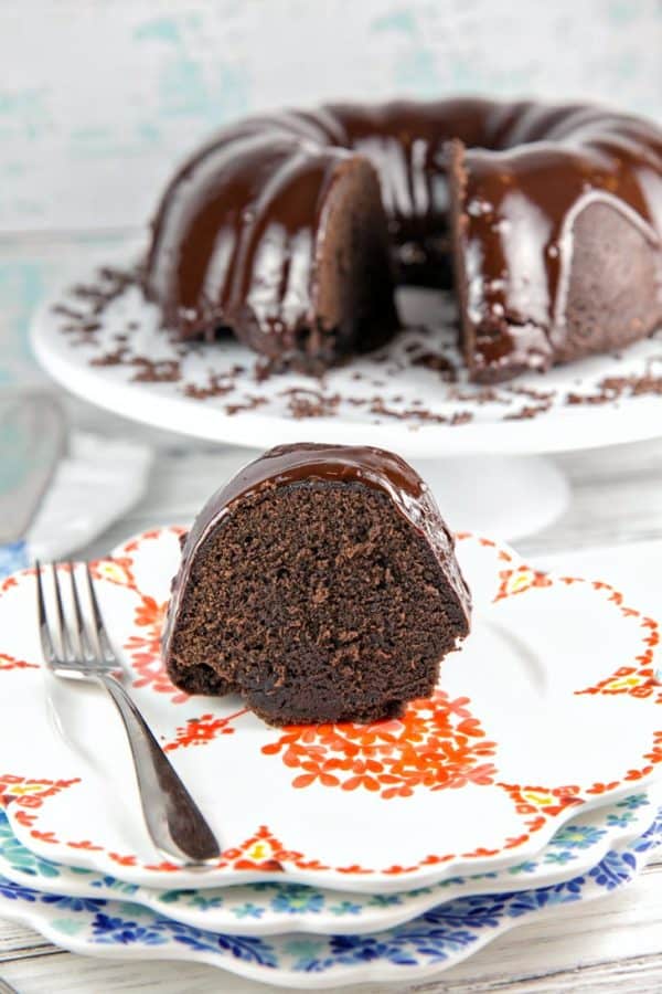 a slice of chocolate fudge bundt cake on a decorating floral dessert plate with the rest of the bundt cake visible in the background