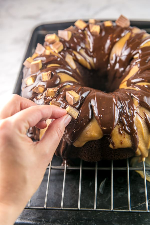 a hand placing chopped peanut butter cups on top of a chocolate bundt cake covered in chocolate and peanut butter ganache