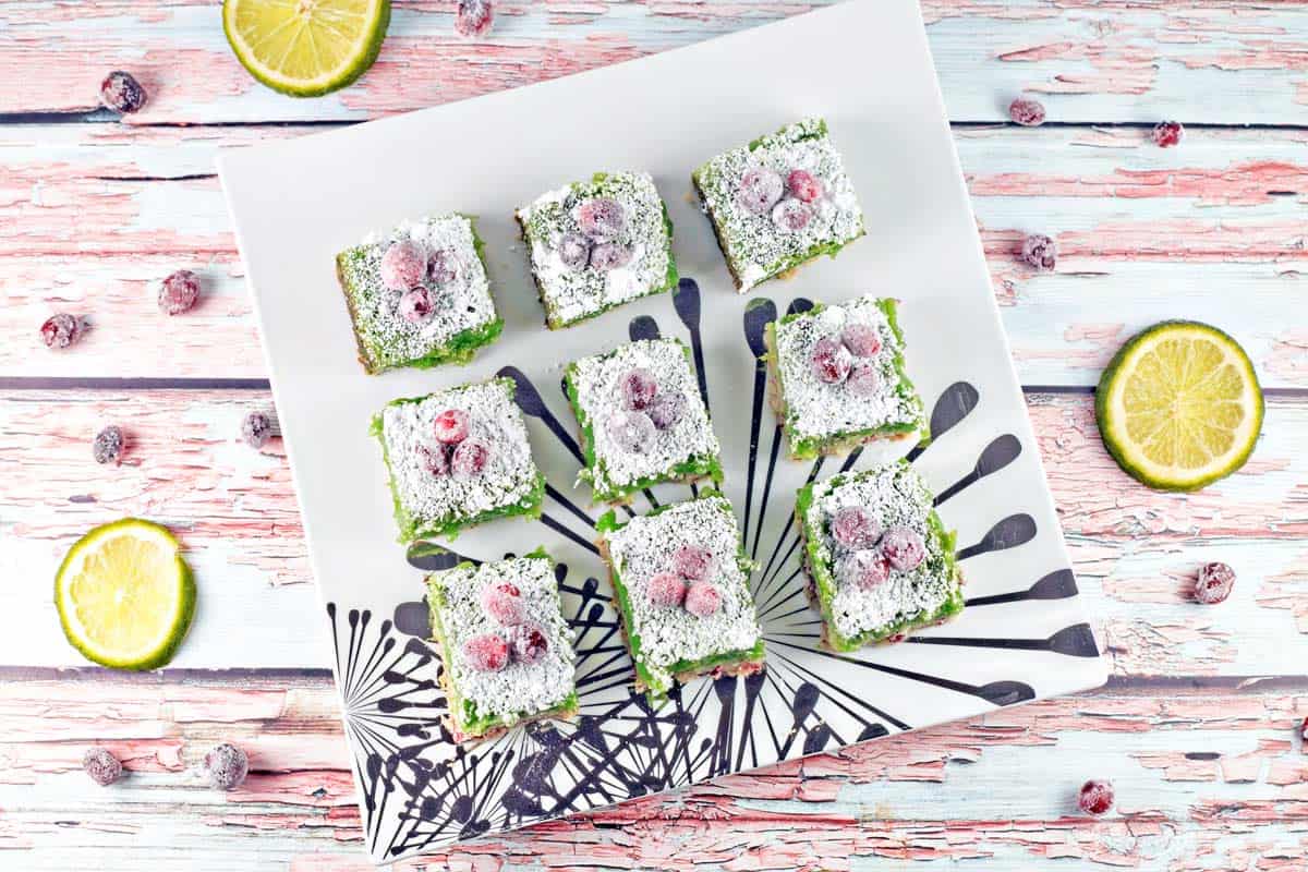 nine cranberry lime bars topped with sugared cranberries on a decorative white plate.