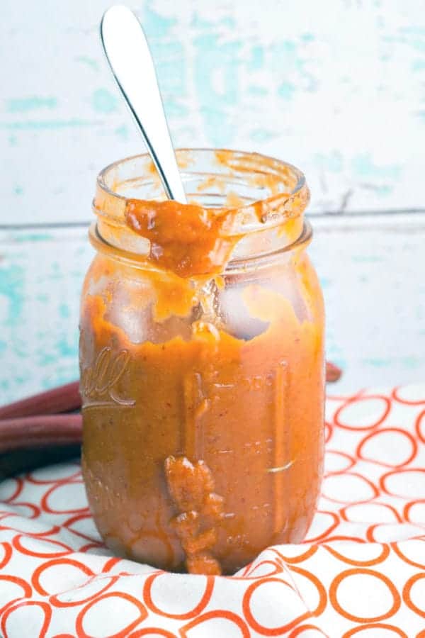 a jar of homemade chipotle rhubarb bbq sauce on a red and white dishtowel against a blue wooden backdrop