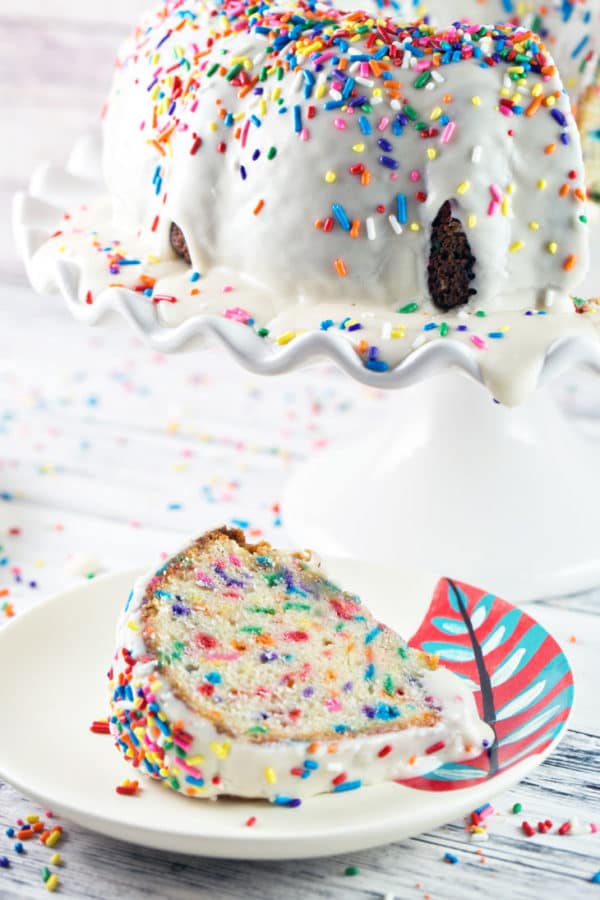 a slice of sprinkle-filled bundt cake on a small plate next to the rest of the uncut cake