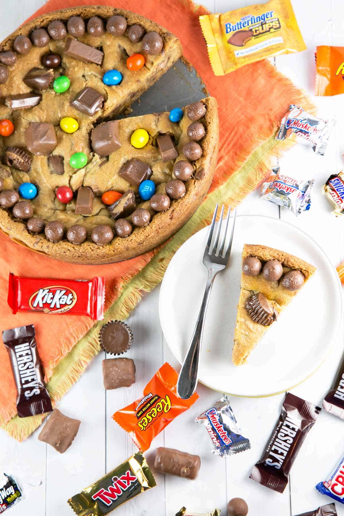 candy cookie cake on an orange placemat with one slice on a white plate surrounded by candy wrappers