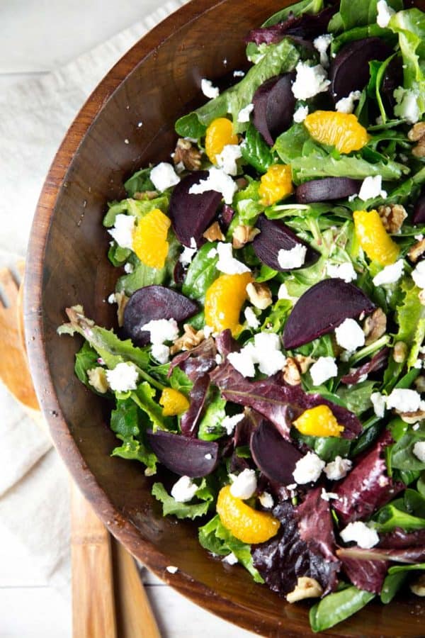 a dark wooden salad bowl filled with leafy greens and topped with roasted beets, mandarin oranges, goat cheese, and candied walnuts