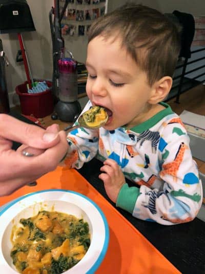 a toddler with his mouth wide open about to eat a big spoonful of sweet potato peanut soup.