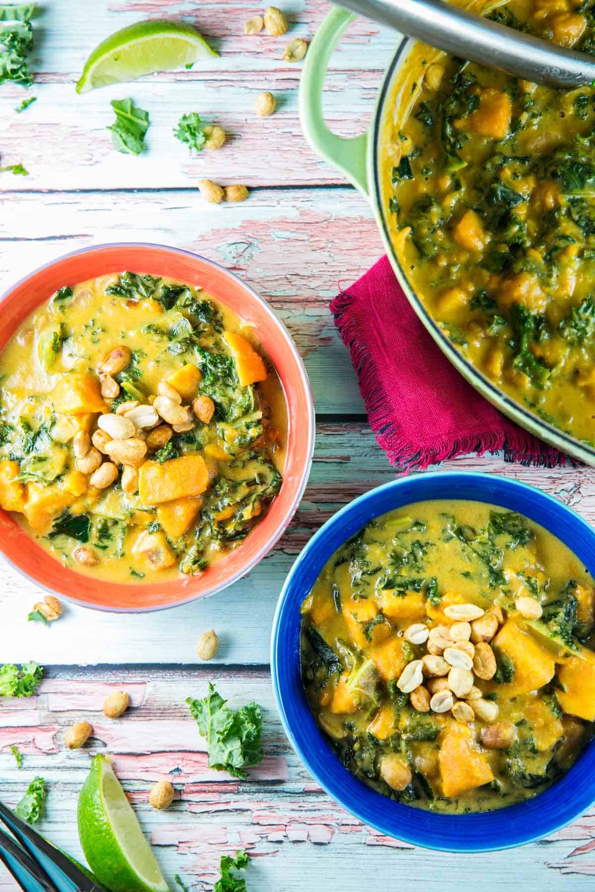 two bowls of african sweet potato soup surrounded by kale and peanuts.