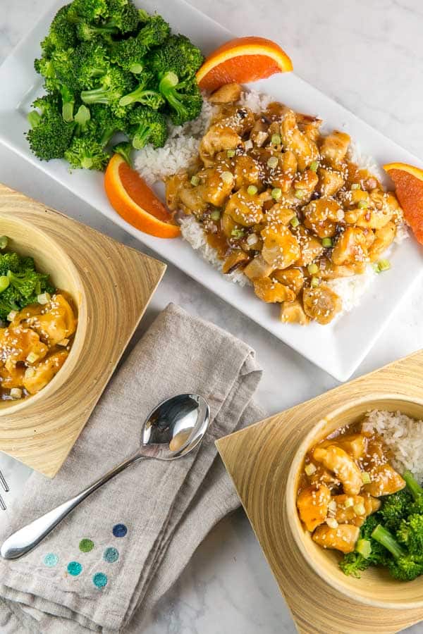 serving dish with rice, orange chicken, and steamed broccoli next to wooden bowls