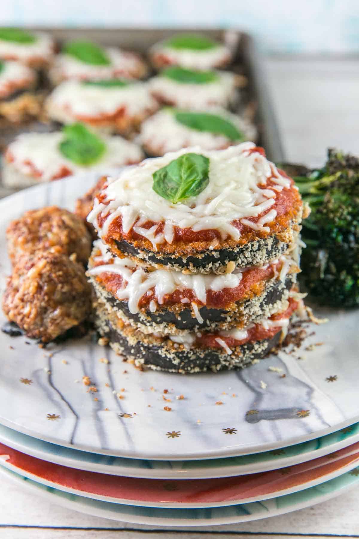 stacks of baked eggplant parmesan on a plate next to meatballs and broccoli.