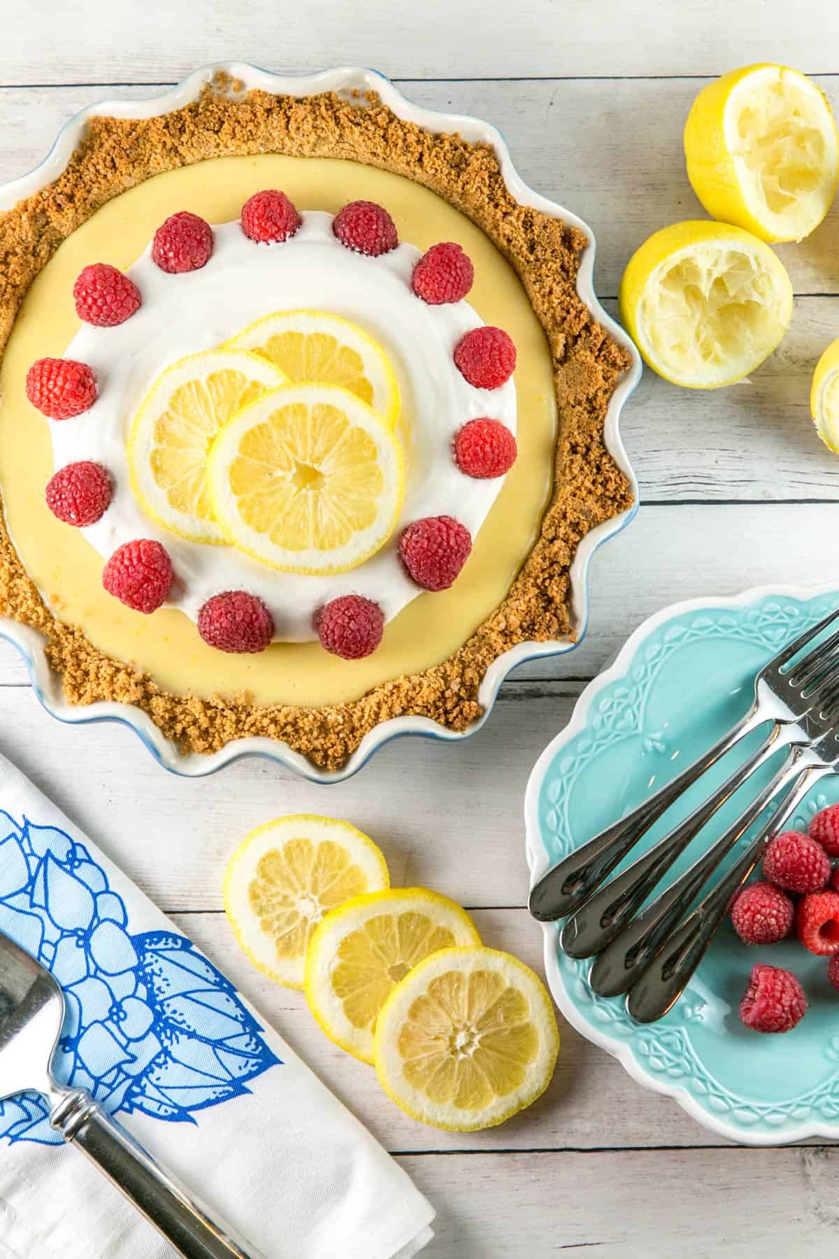lemon custard pie covered in whipped cream and raspberries next to blue dessert plates