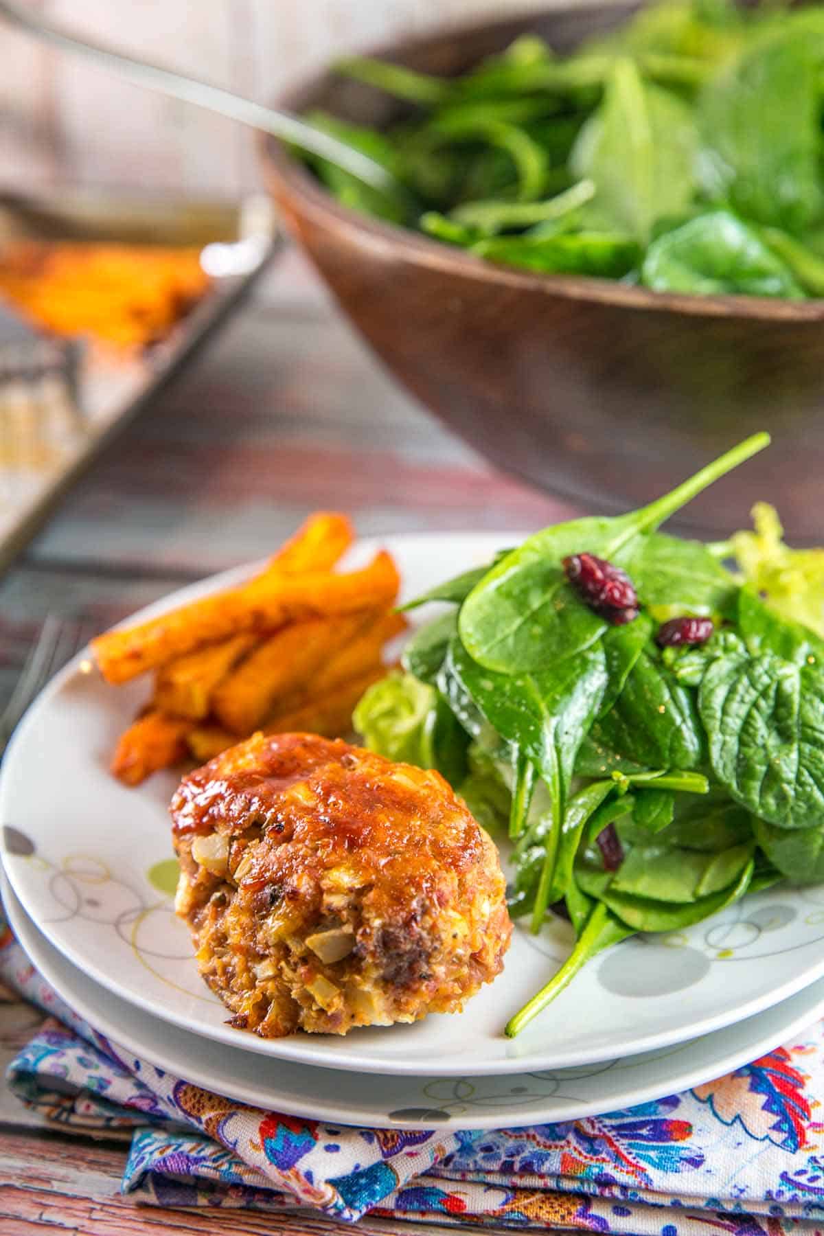 individual mini meatloaf on a dinner plate next to sweet potato fries and a salad.