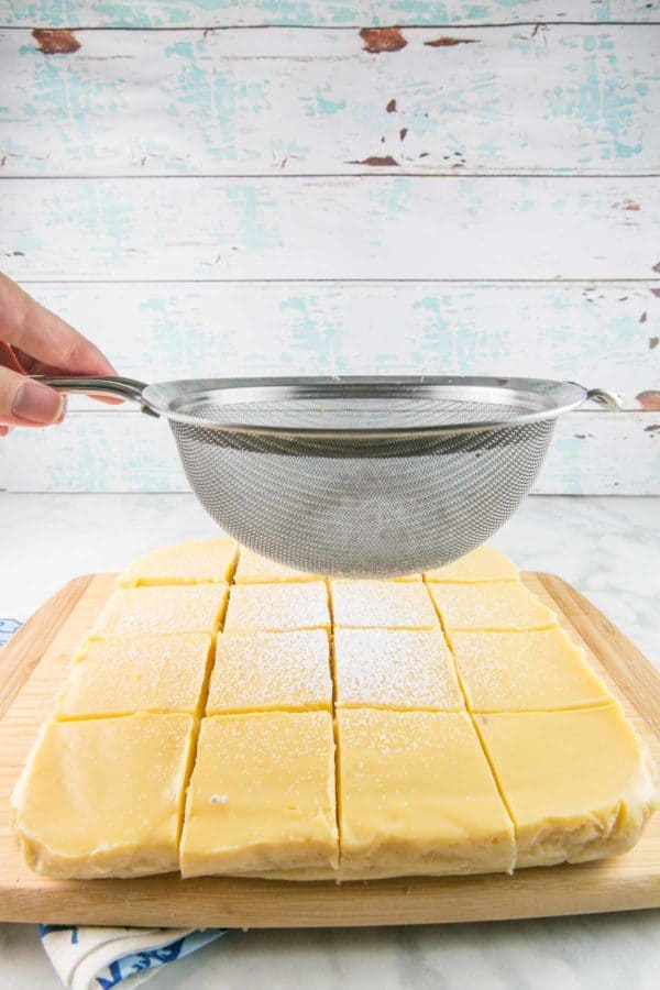 a hand dusting powdered sugar from a mesh sieve on top of lemon pie bars