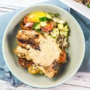 a bowl full of tomato cucumber salad with grilled chicken covered in tahini sauce