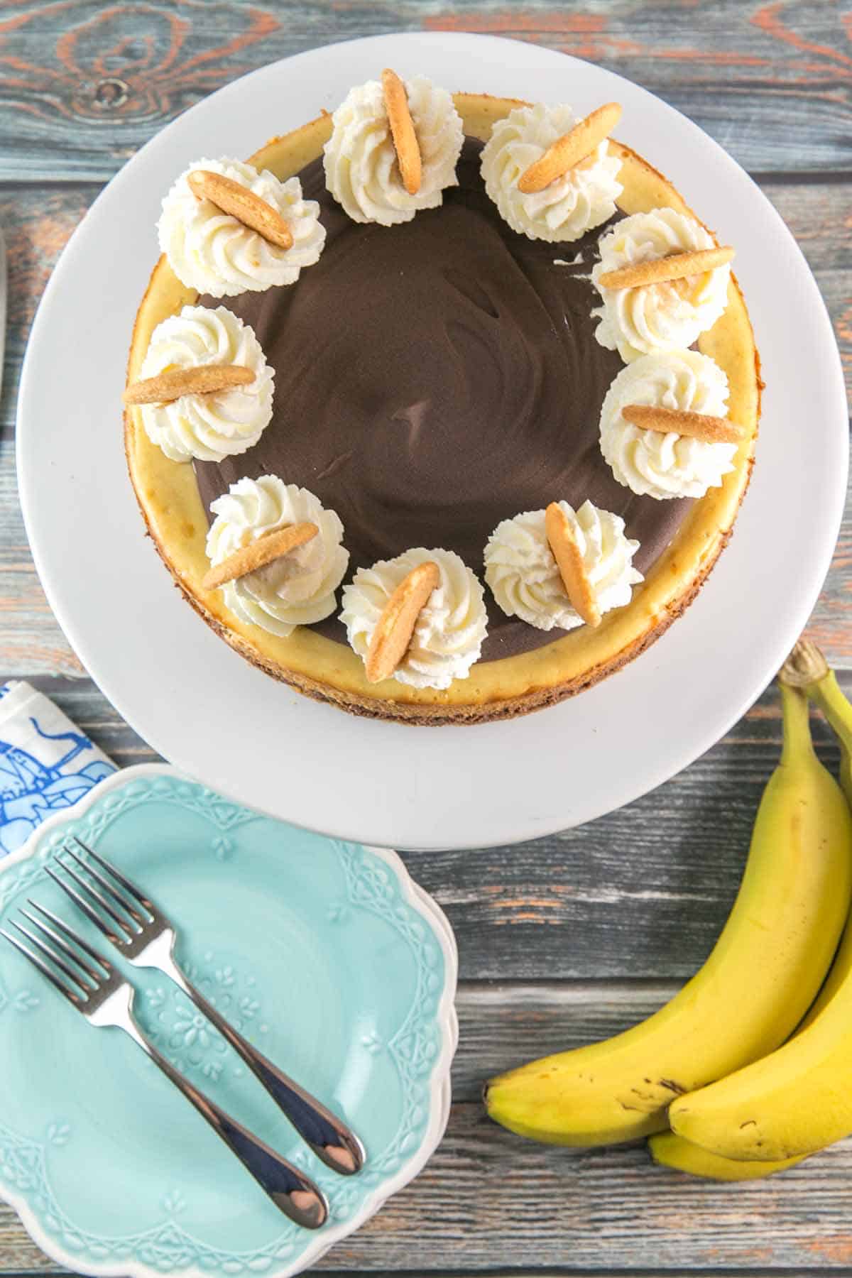 chocolate banana cheesecake covered with ganache and topped with dollops of whipped cream and nilla wafers on a white cake stand