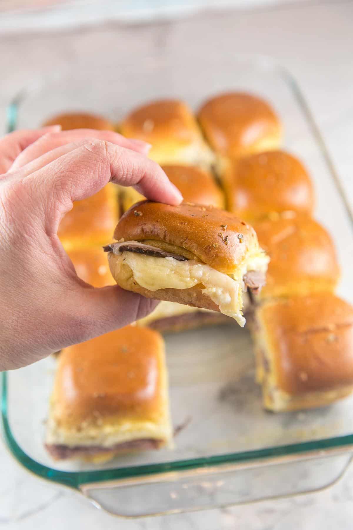 a hand holding a baked roast beef slider with the melted cheese visible along the side of the sandwich