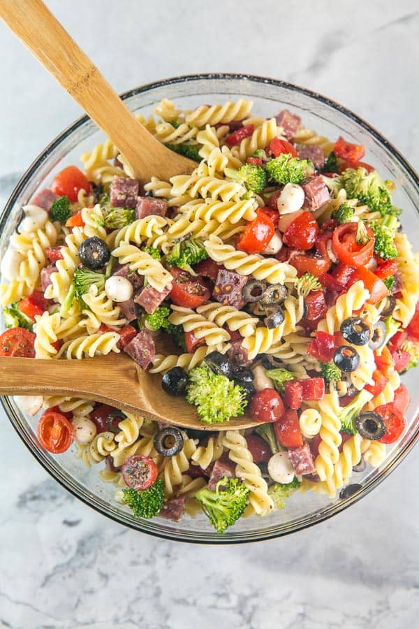 a large glass mixing bowl filled with colorful italian pasta salad and two wooden salad servers