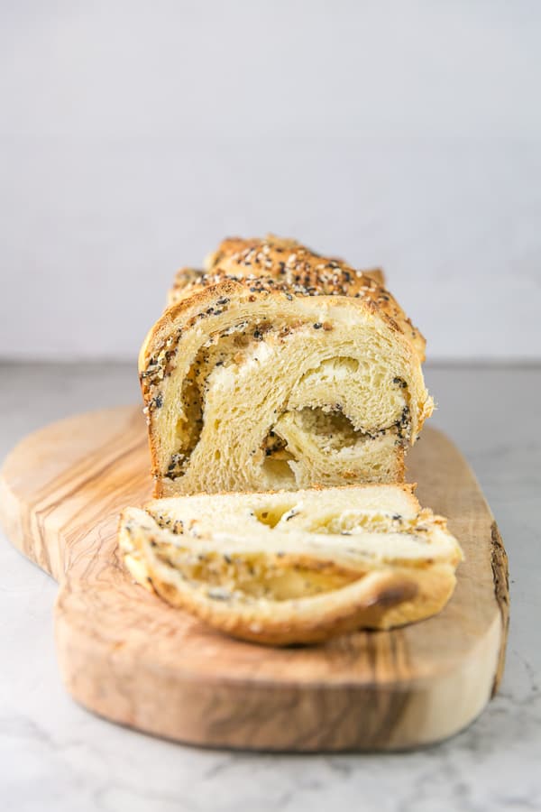 a loaf of everything bagel babka on a wooden board with one slice removed showing the everything bagel seasoning swirls