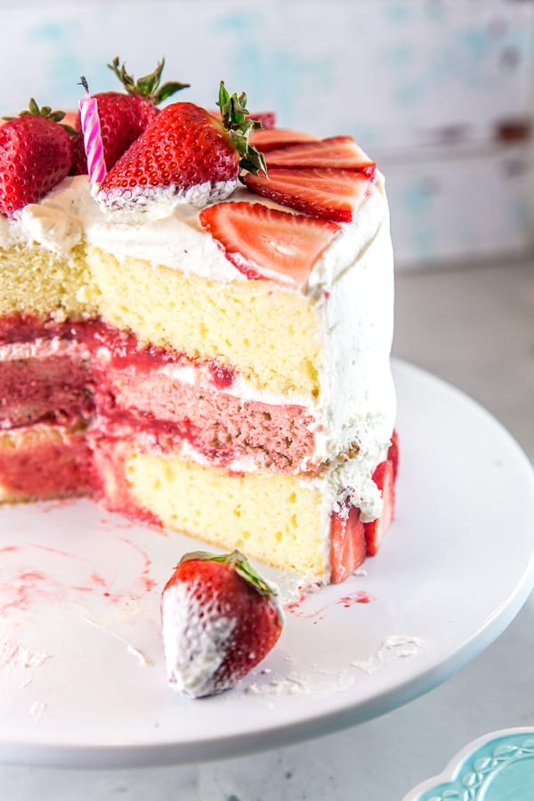 a strawberry layer cake with whipped cream frosting with a large slice of cake removed to show the three distinct layers and strawberry puree filling