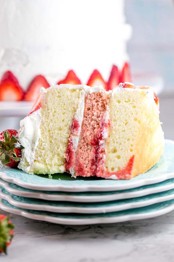 one slice of three layer strawberry layer cake on its side on a stack of blue dessert plates with the rest of the cake in the background