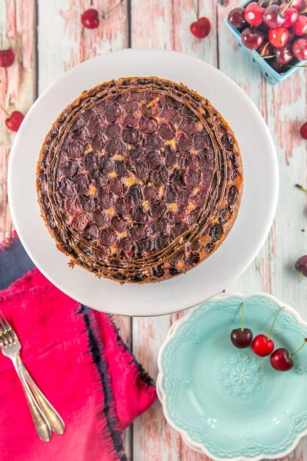 a round chocolate chip cherry upside down cake with circles of cherries on a round white cake plate covered in circles of chocolate drizzle