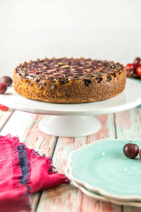 side view of a chocolate chip cherry upside down cake on a white cake stand with fresh cherries spread across the background