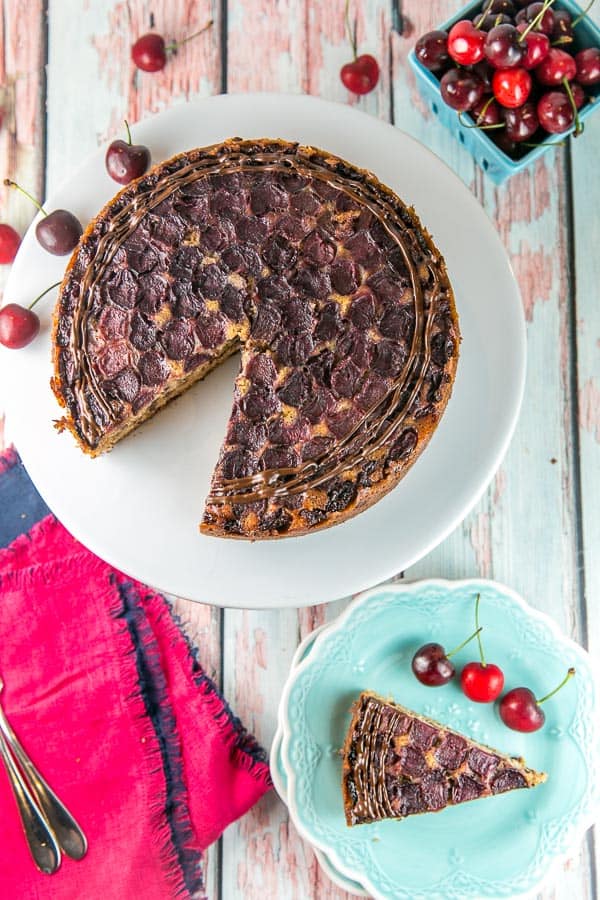 overhead view of a chocolate chip cherry upside down cake on a white cake plate with one slice removed on a blue dessert plate