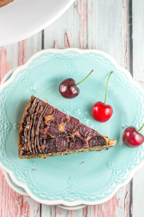 one slice of a chocolate chip cherry upside down cake on a bright blue dessert plate with three fresh cherries a decoration