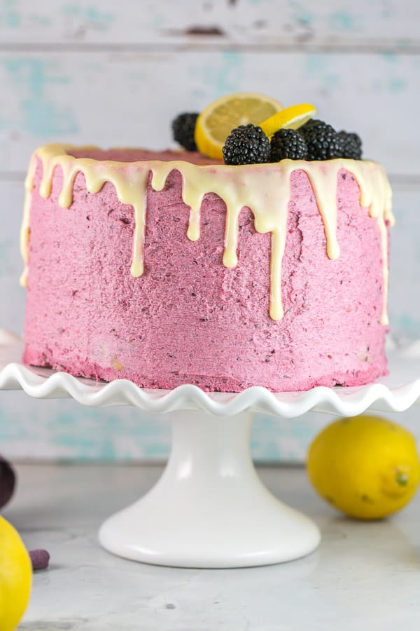 an uncut lemon curd cake covered with pink blackberry buttercream frosting on a white scalloped cake stand