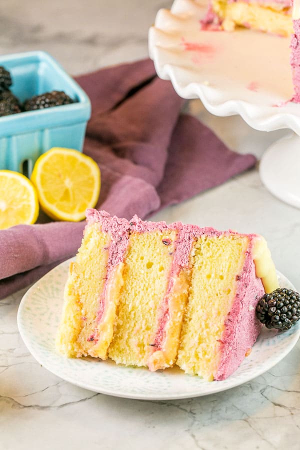a slice of triple layer lemon curd cake with blackberry buttercream on its side with fresh lemons and blackberries visible in the background