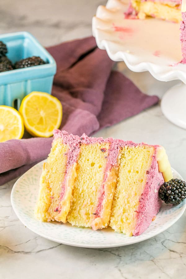 a slice of triple layer lemon curd cake with blackberry buttercream on its side with fresh lemons and blackberries visible in the background