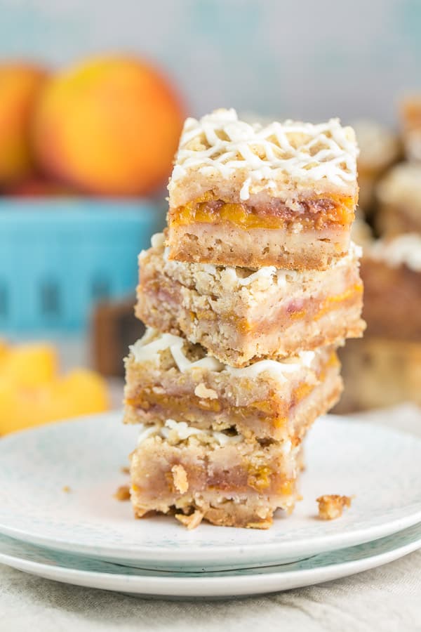 four peach pie bars with oatmeal crumble stacked on top of each other in front of a basket of ripe peaches