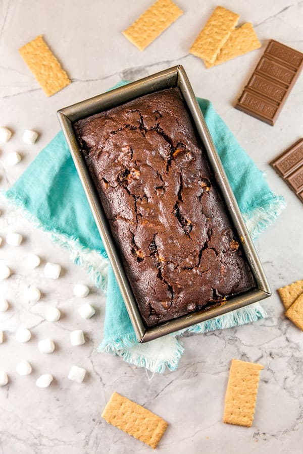 an overhead view of a dark chocolate graham cracker quick bread in a loaf pan surrounded by graham crackers, miniature marshmallows, and a chocolate bar