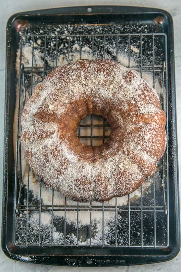 an apple cider donut cake cooling on a rack set on top of a cookie sheet to catch the excess cinnamon sugar sprinkled on the cake