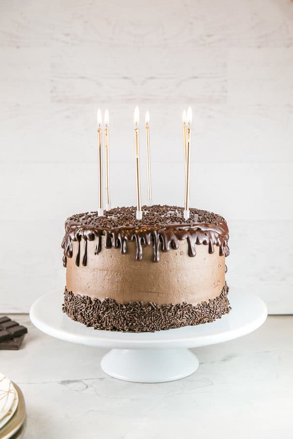 an uncut chocolate layer cake with chocolate ganache and chocolate sprinkles on a white cake stand with lit tall gold pillar candles