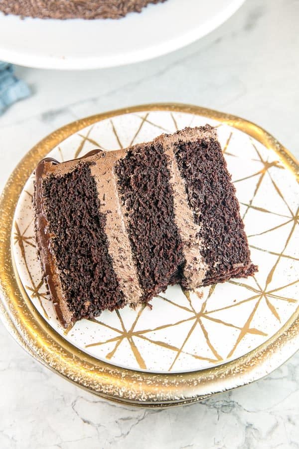 a slice of chocolate layer cake on a white and gold dessert plate showing three layers of dark chocolate cake with thick layers of fluffy chocolate buttercream frosting