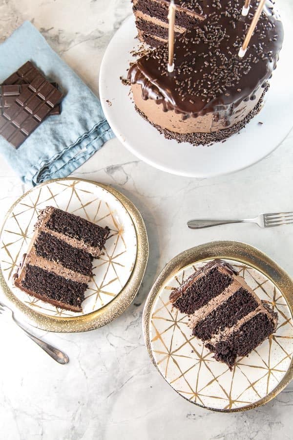 overhead photograph showing two slices of chocolate layer cake with three layers of dark chocolate cake and thick layers of chocolate buttercream on gold dessert plates with the rest of the cake on a white cake stand with tall gold candles in the cake