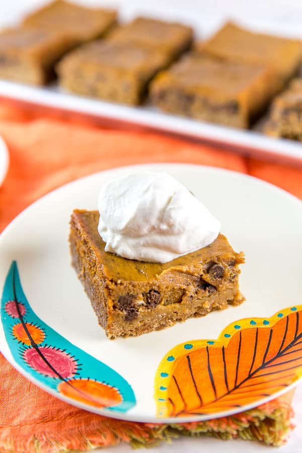 a chocolate chip pumpkin pie bar with a dollop of whipped cream on a fall themed dessert plate sitting on an orange cloth placemat