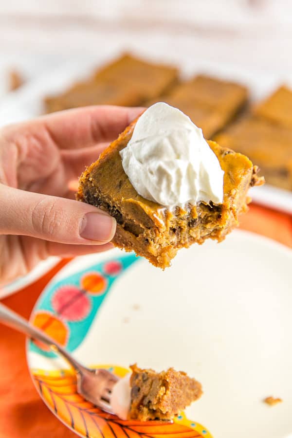 chocolate chip pumpkin pie bar with graham cracker crust and a dollop of whipped cream on top held in a hand with one large bite taken out of the corner