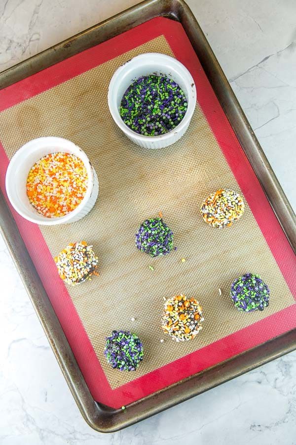 balls of chocolate cooke dough rolled in sprinkles with bowls filled with two different colors of halloween themed sprinkle blends