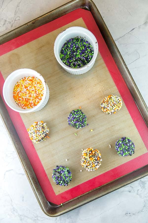 balls of chocolate cooke dough rolled in sprinkles with bowls filled with two different colors of sprinkles.